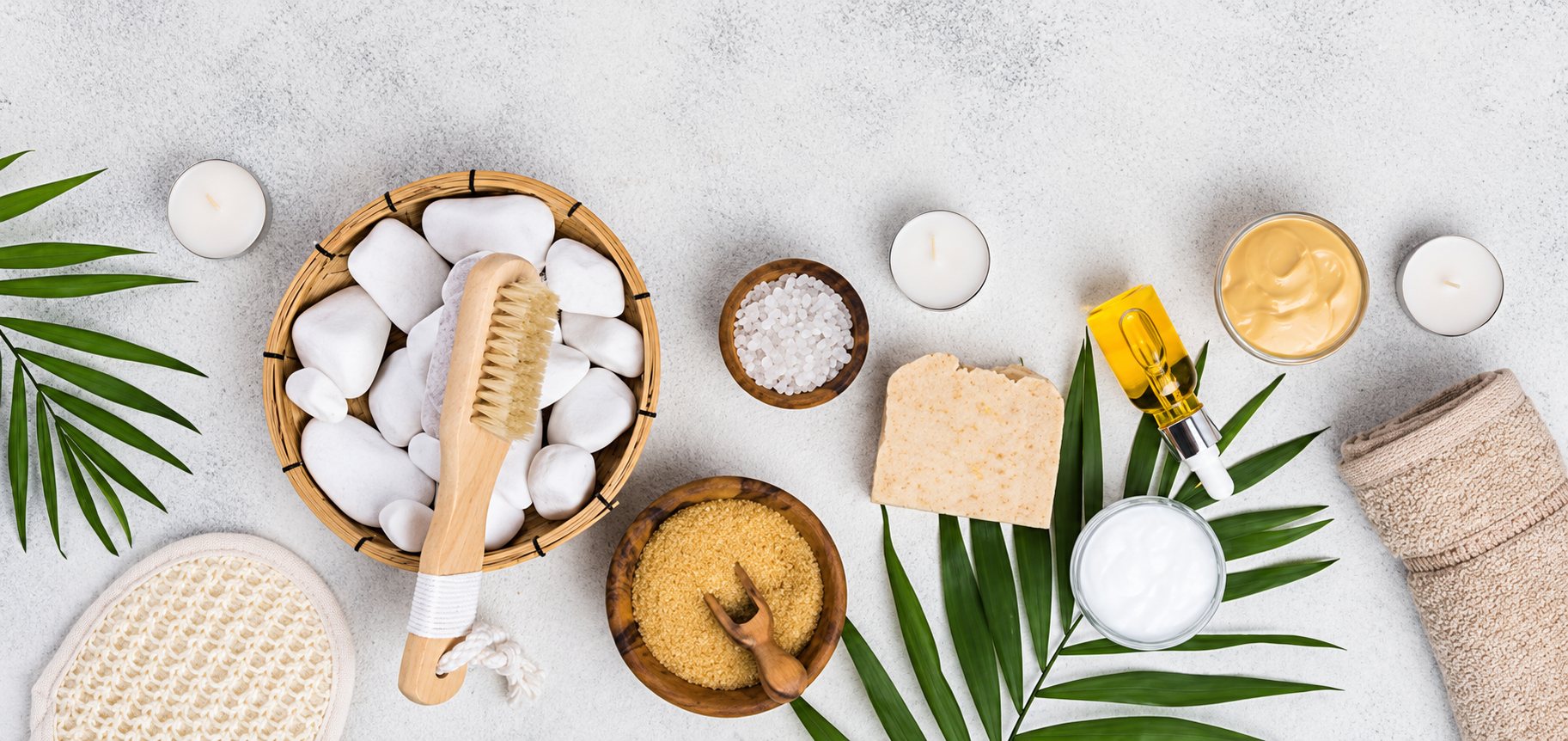 Vue du dessus d'un assortiment de produits de spa : galets blancs dans un panier, brosse en bois, savon, sels, gommage, bougies, serviette et feuilles de palmier.