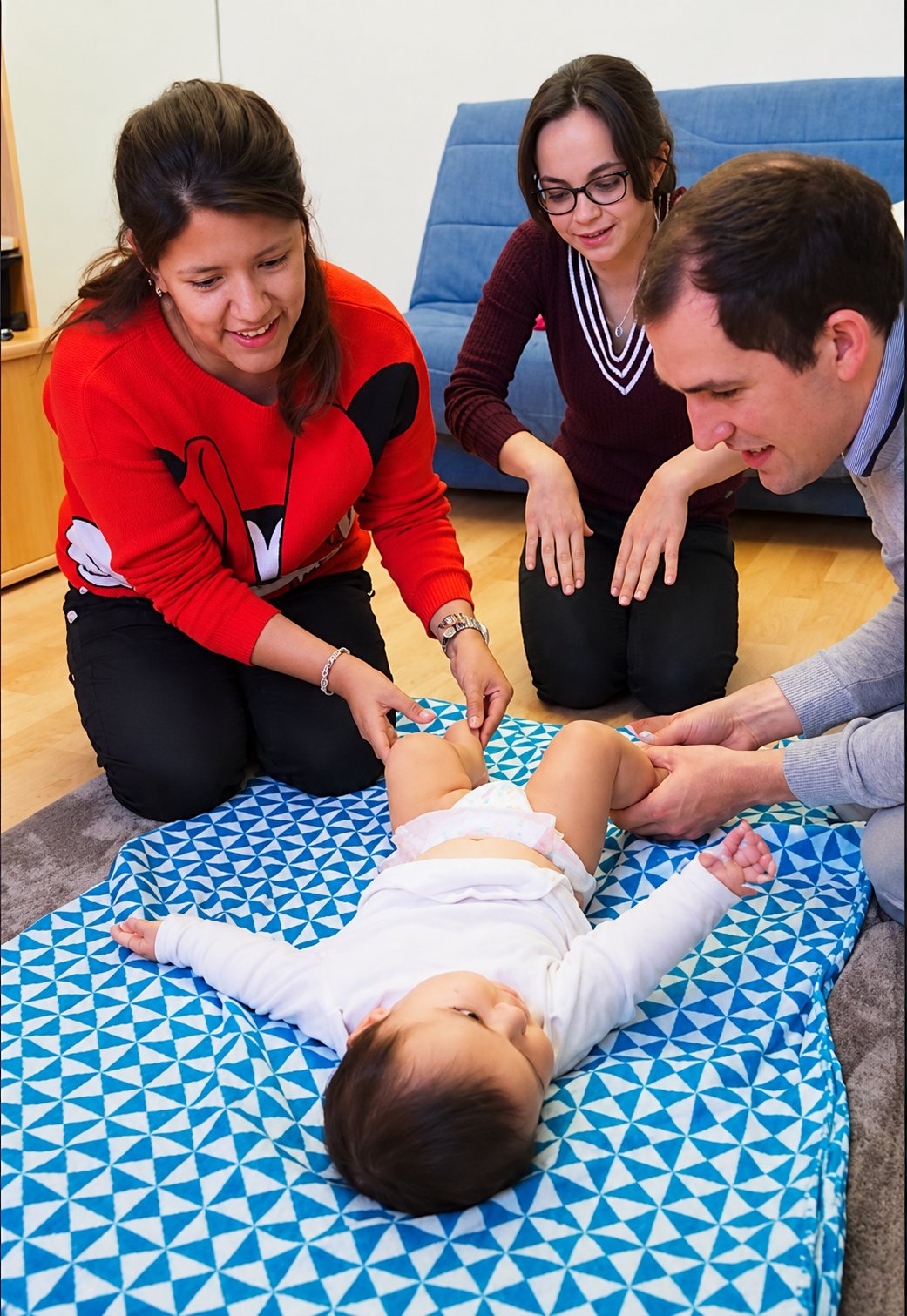 Deux femmes et un homme agenouillés dans un salon observent et touchent un bébé allongé sur un tapis à motifs bleus et blancs lors d'un atelier.