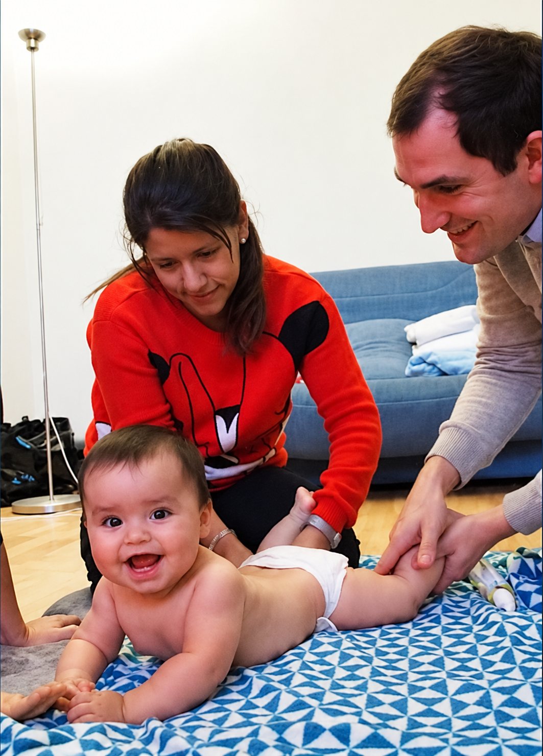 Un bébé souriant est allongé sur une couverture à motifs bleus et blancs tandis qu'une femme en pull rouge et un homme lui massent doucement les jambes.