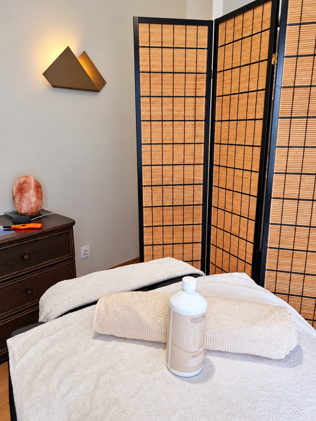 Une cabine de massage avec une table recouverte de serviettes blanches et un flacon d'huile, devant un paravent en bambou et une lampe de sel sur une commode.