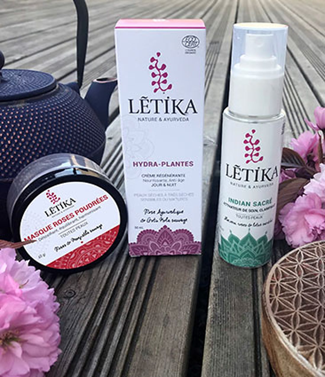 Trois produits de soin Lētika, un masque en pot, une crème en boîte et un flacon pompe, posés sur une terrasse en bois près d'une théière en fonte et de fleurs roses.