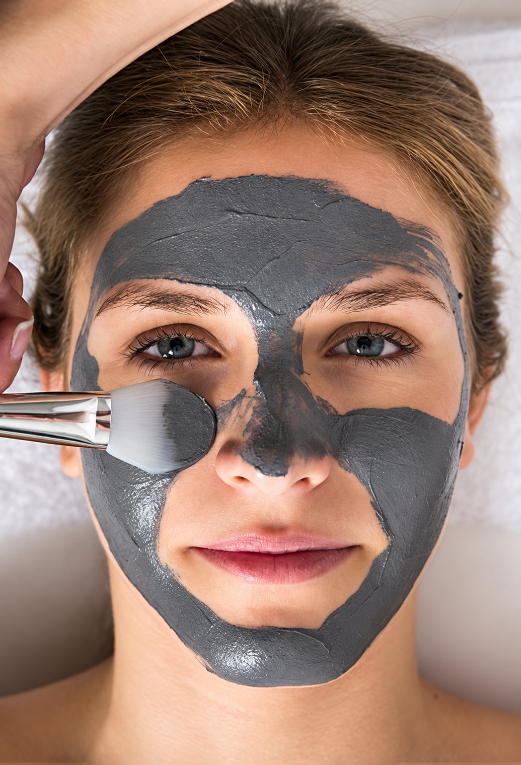 Une femme allongée reçoit l'application d'un masque d'argile grise sur le visage à l'aide d'un pinceau lors d'un soin esthétique.