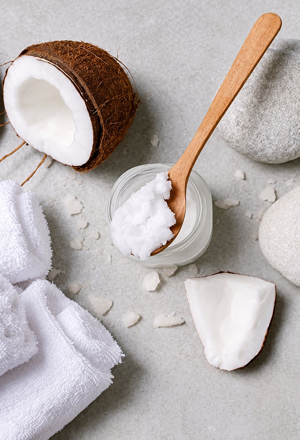 Un pot de verre rempli d'huile de coco avec une cuillère en bois, entouré d'une demi-noix de coco, de galets blancs et de serviettes blanches pliées.