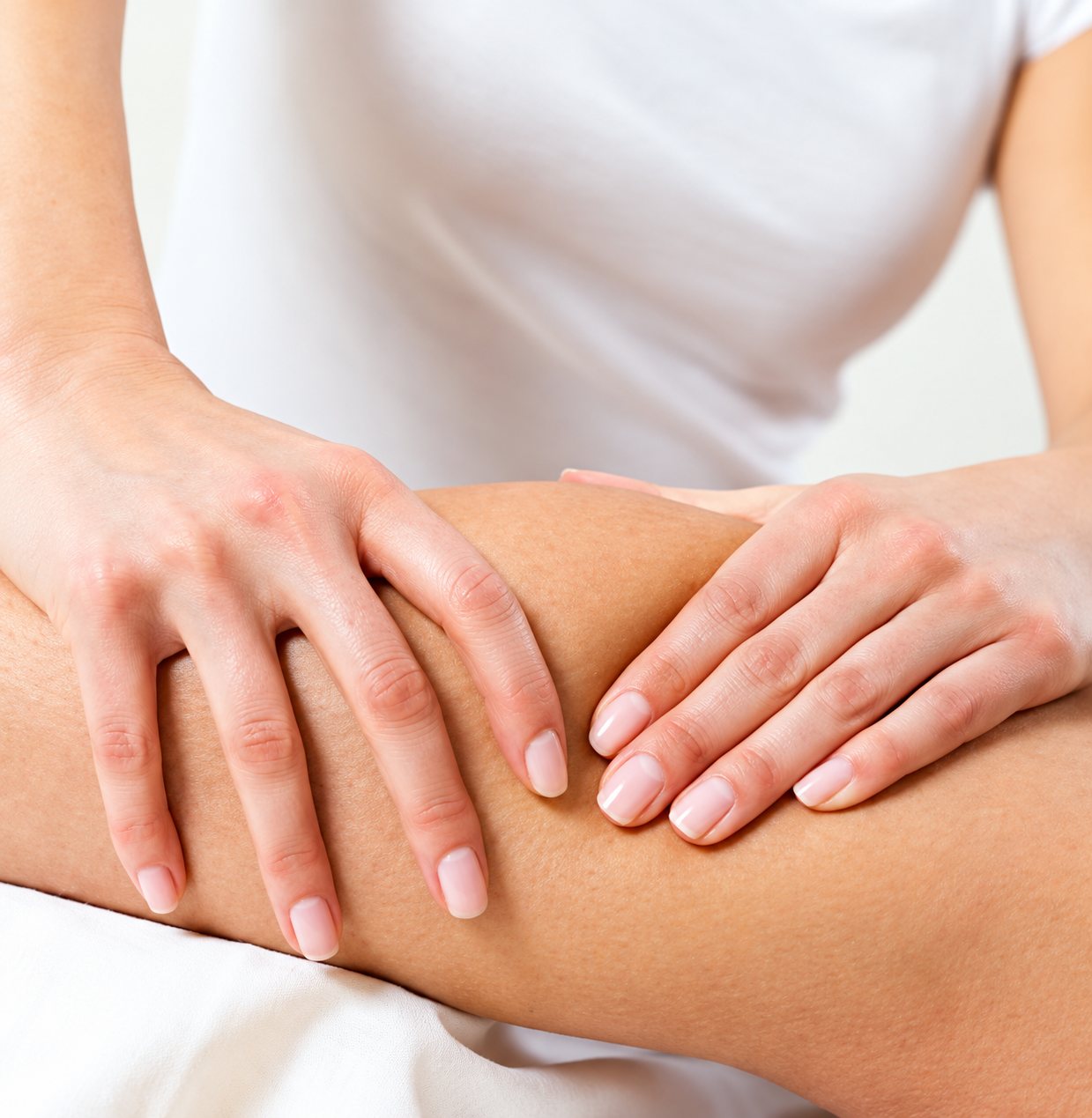 Gros plan sur les mains d'une praticienne en blouse blanche qui masse le mollet d'une personne allongée.
