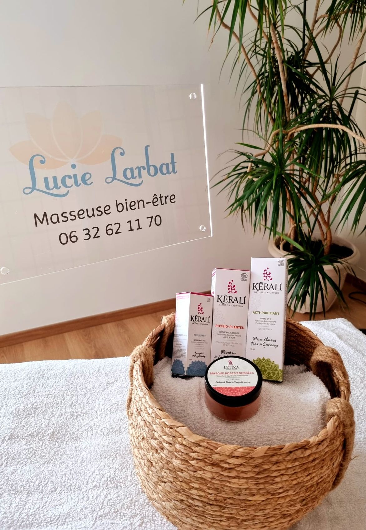 Le panneau transparent « Lucie Larbat, masseuse bien-être » avec son numéro de téléphone, derrière un panier de produits Kérali et une grande plante verte.