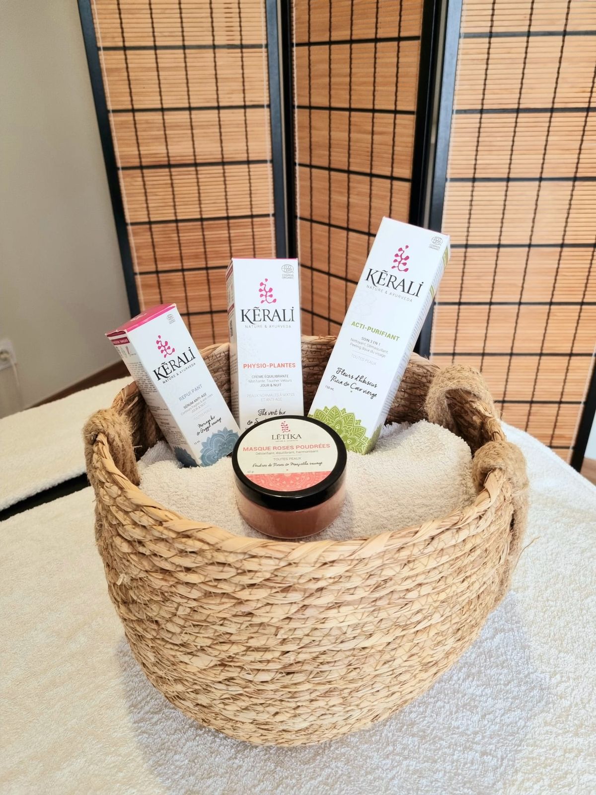 Un panier en paille garni de produits de soin Kérali et d’un pot de masque Lētika, posé sur une serviette blanche devant un paravent en bambou.